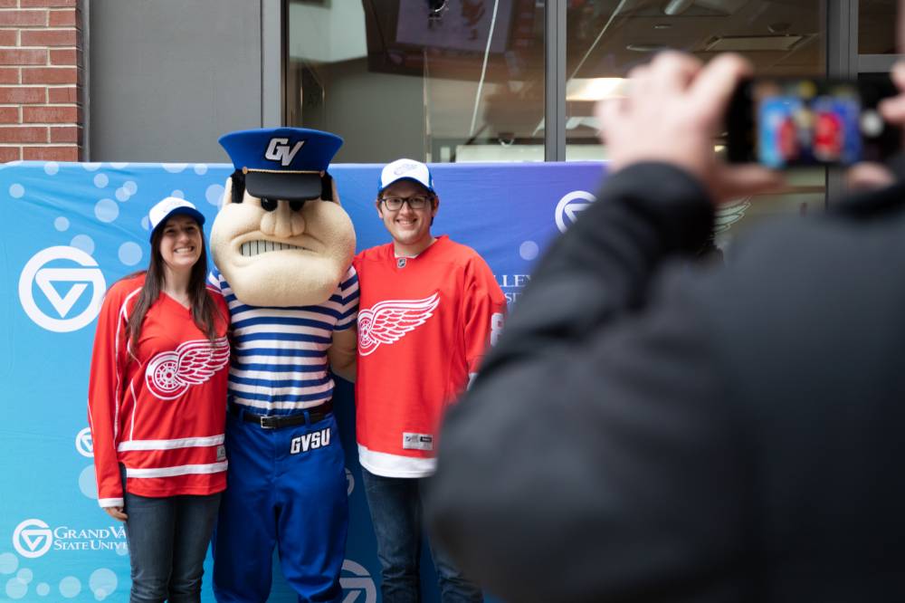 Two alumni, wearing Red Wings Jerseys pose with Louie the Laker at the Detroit Red Wings GVSU Night